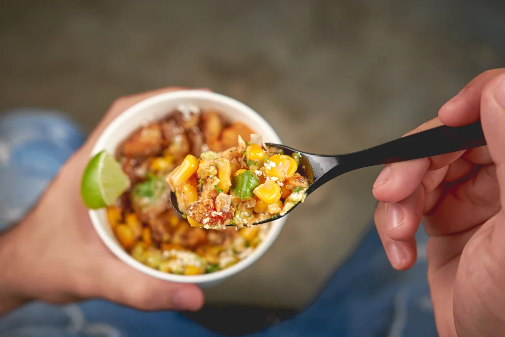 Hand holding spoonful of colorful elote salad. served in a glass on street